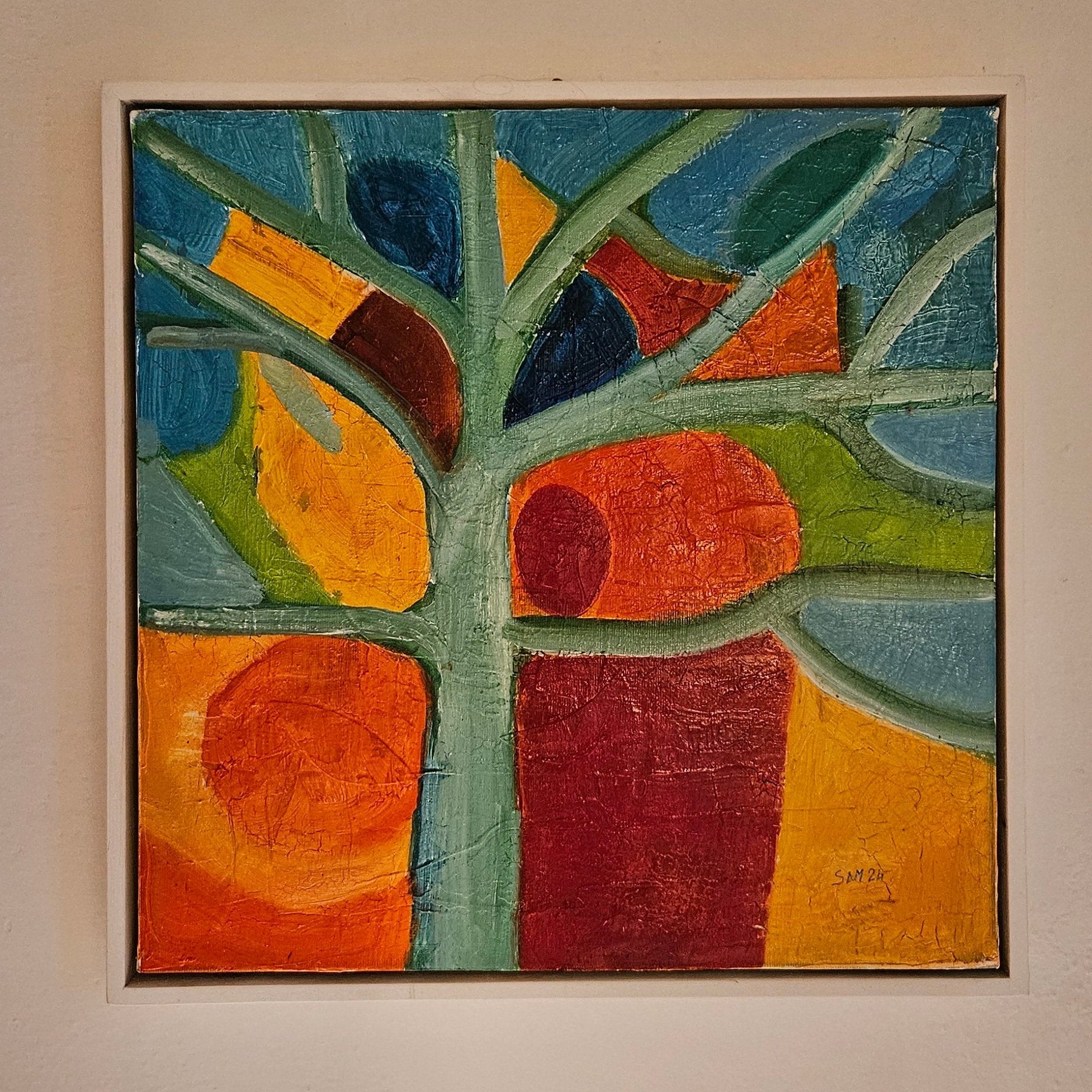 White framed abstract oil painting with green tree with blue, orange red and yellow shapes in the background