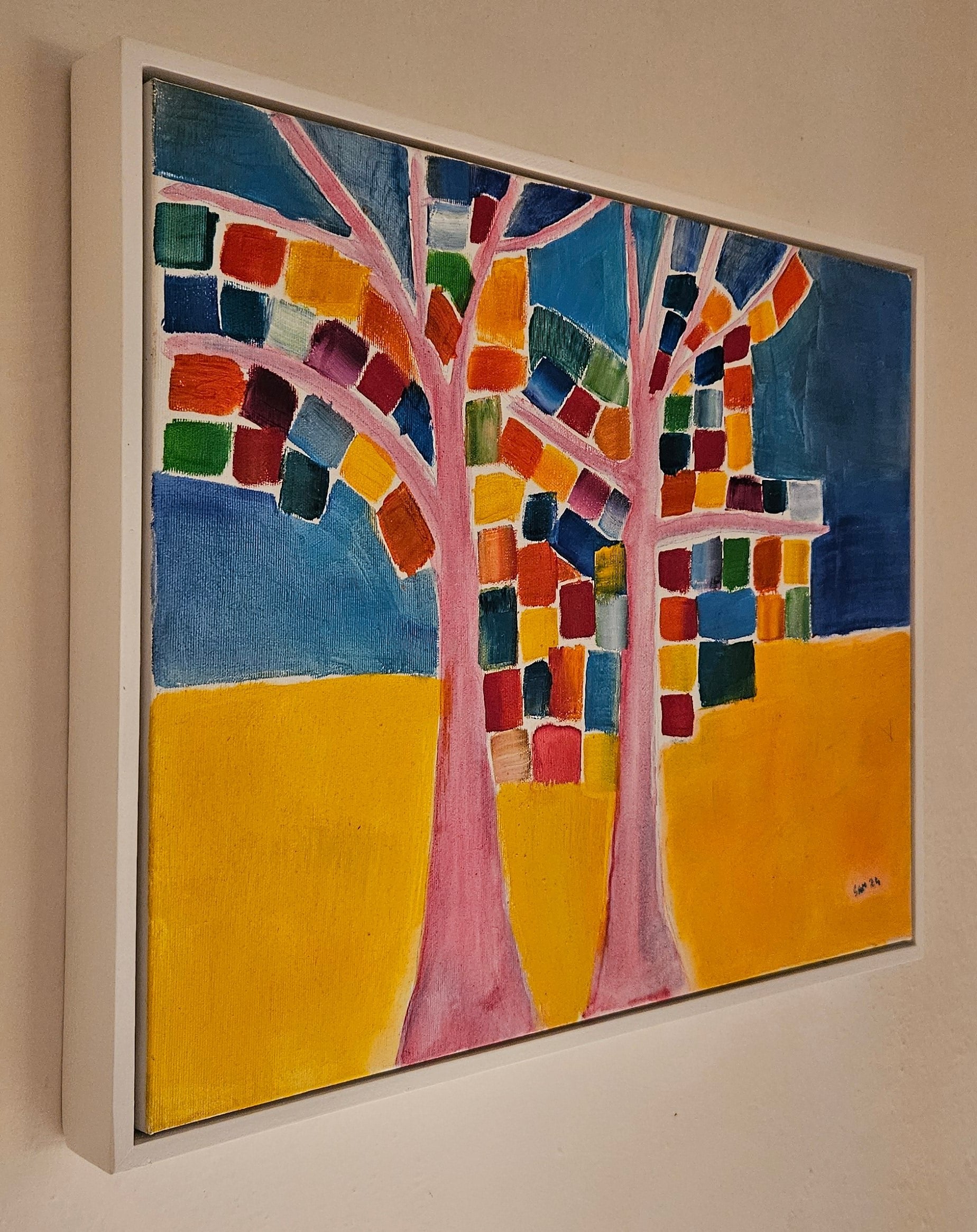 Angled photo of a framed abstract tree oil painting on canvas with white frame. Blue and yellow background with colorful leaves painted as squares.